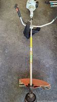 Full-length view of weed wacker laid on concrete floor showing engine housing, straight shaft, handle and cutting head
