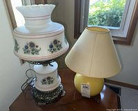 Two table lamps together on wooden table by window. Larger lamp with floral milk glass and metal base, smaller yellow lamp with fabric shade.