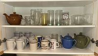 Shelf with teapots, coffee mugs, and glassware including tall glasses and small bowls in a white shelving unit.