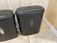 Photo showing the two black shredders side by side on a tile floor, one taller with the clear waste bin window visible.