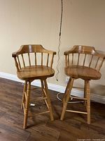 Two wooden bar stools side by side, showing full height, backrests with spindle supports, armrests, and four legs with horizontal support bars.