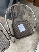 Close-up photo of one brown woven plastic wicker-style chair with tubular metal frame and armrests, showing woven seat and back with diamond pattern.