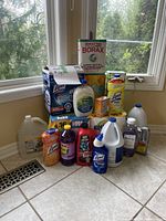 Photo of assorted assorted cleaning products including Borax, Lysol disinfectant wipes, bleach gallons, Drano, Downy, and more on tiled floor near window