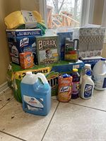 Various household cleaning products and related supplies grouped on floor near window. Products include Lysol wipes, Borax box, bleach bottles, detergent jugs, scrubbies, microfiber towels, tissues.