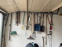 Image shows a variety of used garden tools hung neatly on a white wall under a garage door or ceiling.