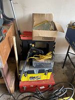 Stack of assorted tools including Stanley toolboxes, a black folding saw horse, a white baseball bat leaning against the shelf, and a cardboard box with papers on top