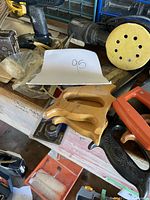 Orbital finishing sander with yellow pad, two wooden hand saws, a staple tool, and black hand saw on wooden surface.