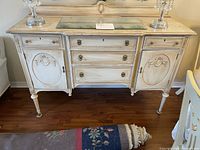Full view of the vintage wooden buffet showing the distressed finish, ornate carved details, glass inset on top, two cabinets with circular raised carvings, and three central drawers with metal handles.