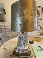 Full view of the lamp showing the gold leaf textured cylindrical lampshade and porcelain base with three female figurines.