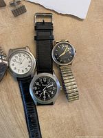Three men's watches with leather and metal bands laid on a table, a pair of cufflinks visible in background.