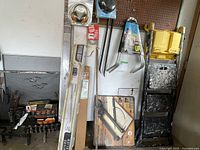 View of various garage items arranged against a wall including concrete trowels, gardening rake, collapsible hand cart, yellow wheelbarrow, and boxed metal braces.