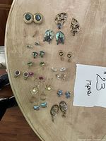 Photo showing 17 pairs of used women's earrings of various styles placed on a wooden surface, featuring designs incorporating abalone shell, leaf motifs, gemstone-like studs, and gold-tone metals.