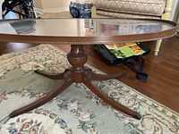 Side view of oval coffee table showing wooden pedestal base and clear glass top on floral rug.