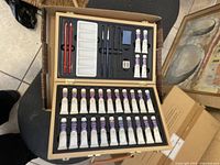 Open wooden paint set case showing 36 paint tubes, paint palettes, brushes, eraser, sharpener, and painting tools arranged inside.