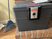 Gray metal safe placed on brick hearth with visible front lock panel marked 'First Alert' and keyhole.