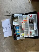 Top view of black plastic fishing tackle box open showing multiple compartments with various fishing tackle items inside, including fishing line, floats, hooks, and bobbers.