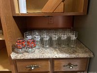 Photo showing 11 clear tall glasses with white Coca-Cola logo and 2 shorter clear glasses with red and black logo arranged on counter.
