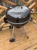 Photograph showing the Weber charcoal kettle BBQ on a three-legged stand with wheels, viewed from the side on a wooden deck.