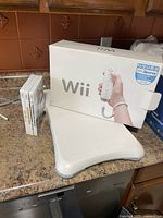 Photo shows Nintendo Wii console, original box, and Wii Balance Board on countertop.