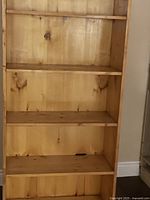 Front view of wooden bookshelf showing five shelves with some wood grain and knots visible, natural wood finish, placed against a wall near a staircase.
