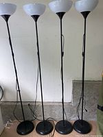 Four black metal uplighter floor lamps with white bowl-shaped shades arranged in a row against a white wall.