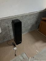 Photo of a black tower-style air purifier standing on the floor against a gray wall and concrete baseboard, with attached electrical cord and plug.