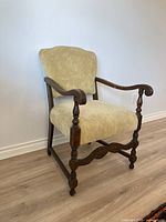 Photo of an antique arm chair showing wooden frame, curved armrests, and light beige fabric upholstery on seat and backrest.