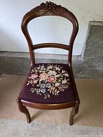 Photo of a wooden antique chair showing its carved backrest and detailed floral embroidery on the seat upholstery.