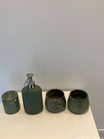 Four green-toned bathroom accessories on a white surface against a plain wall, including soap dispenser, toothbrush holder, and two small containers.