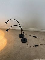 Photo of two black bedside lamps with adjustable gooseneck arms placed on the floor against a white wall, showing the base, cords, and inline switches, demonstrating their overall condition and style.