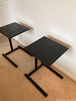 Pair of black metal bedside tables with square tops and tubular C-shaped bases seen from above on a beige carpet against a white wall.