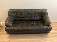 Black leather three-person sofa front view showing cushioned armrests and padded backrest on light wood floor.