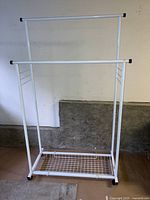 Front view of white metal clothing rack with two horizontal hanging rails, wire mesh lower shelf, and black plastic end caps on rails. Wheels visible on bottom.