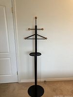 Single photo of the black metal clothing stand with wooden knobs, round base, and a small round tray mid-way. The background is a plain wall with a white door to the side.