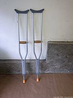 Photo showing two adjustable aluminum crutches standing upright against a white wall with a concrete ledge behind.