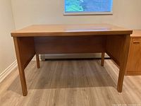Front-facing image of the custom solid wood desk showing the full rectangular tabletop and four sturdy legs on wood grain flooring against a plain wall with window above.