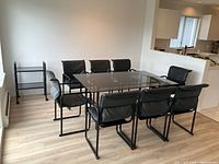 Wide angle photo showing the rectangular glass dining table with eight black padded chairs arranged around it, next to a window.