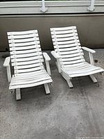 Pair of white wooden adjustable deck chairs with slatted seats and backs shown outdoors on concrete patio.