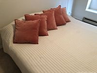 Photo of the king-size bed with beige/cream quilted cover and six terracotta decorative pillows lined against the headboard. Bed frame visible on sides in beige/cream color.