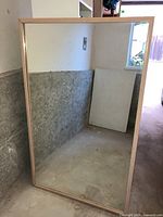 Photo shows a large rectangular mirror leaning against a wall in an unfinished space. The frame is made of light natural wood and the mirror surface is clear, reflecting the concrete floor and walls.