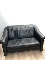Front view of a black leather love seat with padded armrests and vertical stitching on the backrest, placed on a wooden floor.