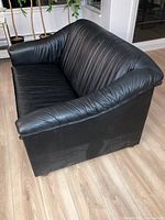 Side view of a black leather loveseat with padded, rolled armrests and backrest on a light wooden floor.