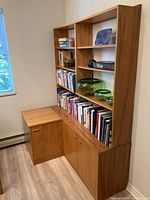 Photo shows two parts of a custom-built New Zealand Kauri wood furniture unit: a bookshelf with five shelves and a bottom double-door storage cabinet, and a separate square cabinet with one door and recessed handle, adjacent to the bookshelf, all holding books and decorative items.