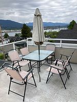 Photo shows outdoor patio dining set with square glass table, beige umbrella closed and six matching beige folding chairs around it. Patio deck, railing, and distant mountainous view visible.
