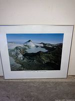 Framed photo showing an aerial view of Crater Lake at Mt. Ruapehu, New Zealand, with mountainous terrain and the lake inside the caldera. The photo is matted and framed in a simple silver or light-colored frame.