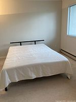 Full view of the queen size bed showing metal headboard, mattress covered with a light sheet, situated in a plain room.