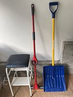 Photo showing a white metal step stool with black cushioned top, a red mop with black grip, and a blue snow shovel with yellow handle and blue D-grip, against a white wall.