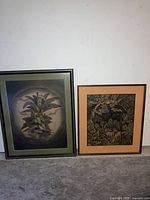 Photo showing two framed Balinese drawings leaning against wall on floor, capturing full view of both artworks and frames.