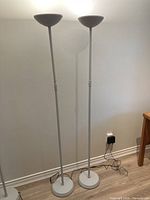 Two white uplighter floor lamps standing side by side against a plain wall, showing full height and style of lamps.