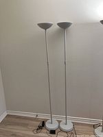 Two white uplighter floor lamps standing side by side against a wall on a wooden floor, showing full height and base details.